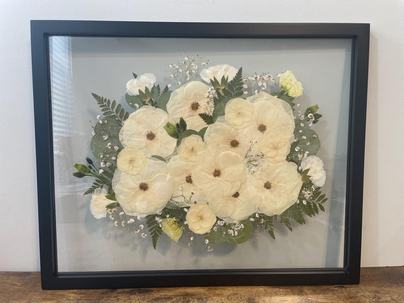 May include: A framed floral arrangement featuring cream-colored flowers with brown centers, surrounded by green foliage and baby's breath. The arrangement is encased in a black rectangular frame with a glass front.