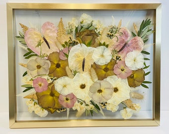 May include: A framed pressed flower arrangement with a variety of flowers in shades of pink, white, and brown. The flowers are arranged in a circular pattern and are surrounded by greenery.
