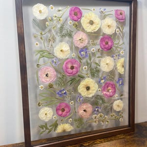 May include: A framed floral arrangement featuring pressed flowers in shades of pink, yellow, and white, with small blue and white accents. The flowers are arranged on a clear background within a dark brown wooden frame. The arrangement is displayed on a wooden surface.