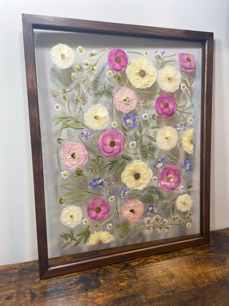 May include: A framed floral arrangement featuring pressed flowers in shades of pink, yellow, and white, with small blue and white accents. The flowers are arranged on a clear background within a dark brown wooden frame. The arrangement is displayed on a wooden surface.