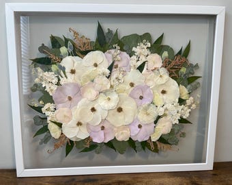 May include: A framed floral arrangement featuring a variety of dried flowers in shades of white, cream, and pale purple. The arrangement includes green leaves and delicate sprigs, all encased in a white frame.