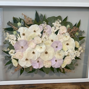 May include: A framed floral arrangement featuring a variety of dried flowers in shades of white, cream, and pale purple. The arrangement includes green leaves and is encased in a white rectangular frame.