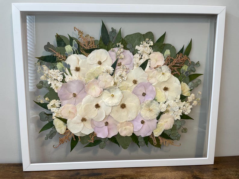 May include: A framed floral arrangement featuring a variety of dried flowers in shades of white, cream, and pale purple. The arrangement includes green leaves and is encased in a white rectangular frame.