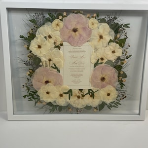 May include: A framed floral arrangement featuring a wreath of cream and pink flowers. The flowers surround a cream-colored card with elegant script. The frame is white and square, creating a clean, modern look. The arrangement is likely for a wedding or special occasion.