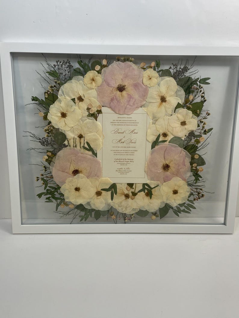 May include: A framed floral arrangement featuring a wreath of cream and pink flowers. The flowers surround a cream-colored card with elegant script. The frame is white and square, creating a clean, modern look. The arrangement is likely for a wedding or special occasion.