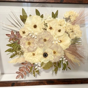 May include: A framed floral arrangement featuring a bouquet of dried flowers in shades of cream, beige, and brown. The arrangement includes various flower types, leaves, and grasses, all set against a white background within a dark wooden frame.