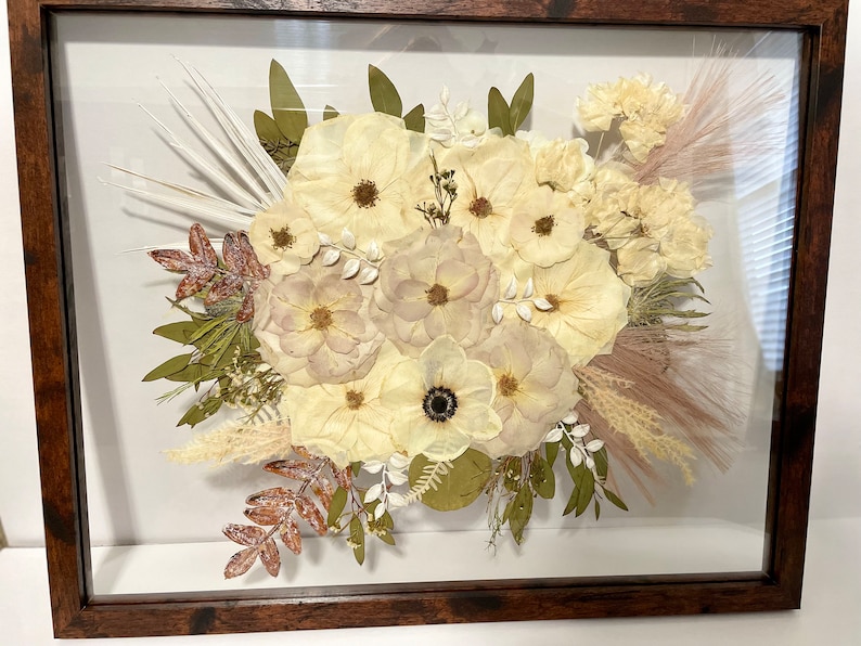 May include: A framed floral arrangement featuring a bouquet of dried flowers in shades of cream, beige, and brown. The arrangement includes various flower types, leaves, and grasses, all set against a white background within a dark wooden frame.