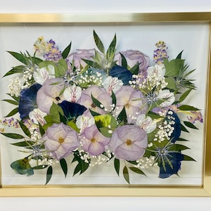 May include: A framed floral arrangement featuring pressed flowers in shades of purple, white, and blue. The flowers are arranged in a bouquet style and encased in a gold-colored rectangular frame. The arrangement includes roses, pansies, and other greenery.