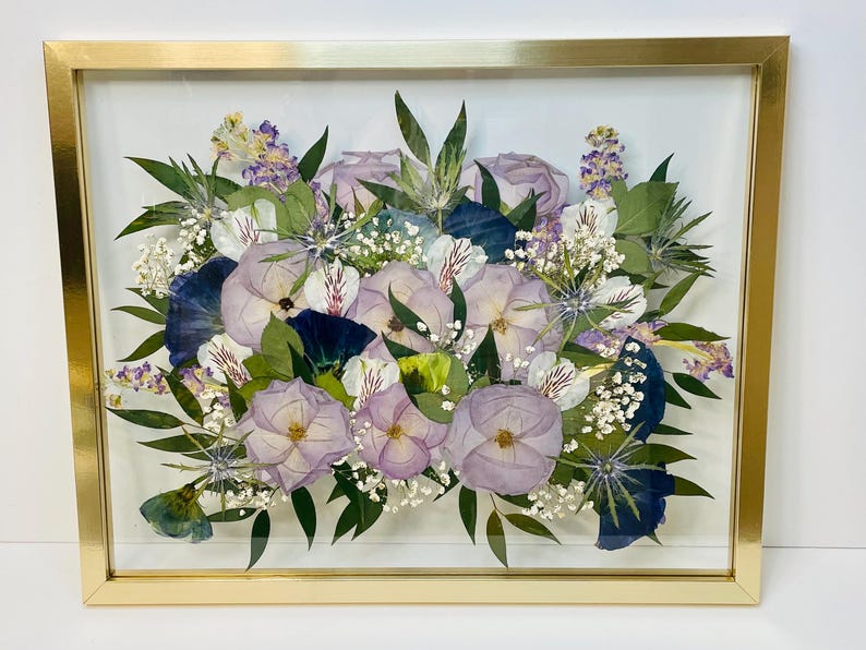 May include: A framed floral arrangement featuring pressed flowers in shades of purple, white, and blue. The flowers are arranged in a bouquet style and encased in a gold-colored rectangular frame. The arrangement includes roses, pansies, and other greenery.