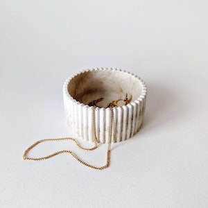 May include: A white ceramic trinket dish with a ribbed design. The dish is filled with gold jewellery, including a necklace and earrings.