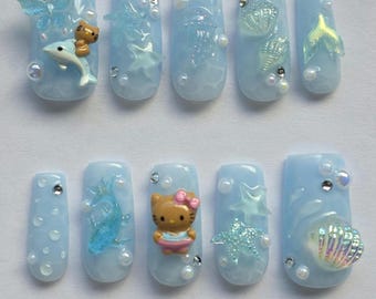 Peut inclure: Ongles à pression bleu clair avec divers ornements sur le thème de l'océan. Les motifs comprennent un papillon, un dauphin, un ours en peluche, une méduse, des coquillages, une queue de sirène et un personnage Hello Kitty. Chaque ongle est orné de perles et de strass.
