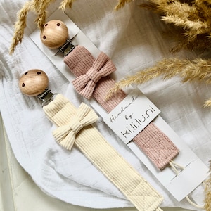 Könnte beinhalten: Zwei handgefertigte Baby-Beißringe mit Holzperlen und einer Schleife. Die Beißringe sind aus einem weichen, strukturierten Stoff gefertigt und in den Farben Rosa und Creme erhältlich. Die Verpackung enthält ein Etikett mit der Aufschrift "Handmade by K.I.I.I.U.N.I."