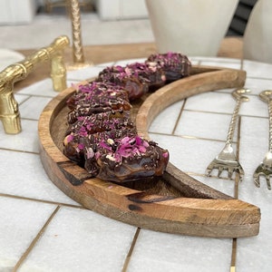 Könnte beinhalten: Ein hölzerner, halbmondförmiger Serviertablett mit dunkler Holzoptik hält eine Reihe von schokoladenüberzogenen Datteln, die mit rosa Rosenblüten verziert sind. Das Tablett befindet sich auf einer weißen Marmorfläche mit goldenen Akzenten.