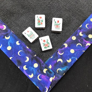 May include: Four white Mahjong tiles with red and green illustrations and Chinese characters are arranged on a black mesh surface. A blue fabric with gold moons and circles borders the tiles.