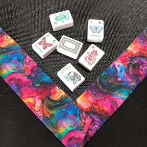 May include: Six white Mahjong tiles featuring various designs including a green dragon, a red dragon, a butterfly labeled SPRING, a butterfly labeled WINTER, a panda labeled JOKER, and a blank tile, resting on a vibrant, multi-colored fabric border.