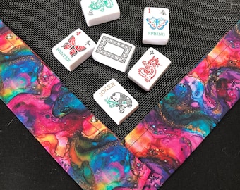May include: Six white Mahjong tiles featuring various designs including a green dragon, a red dragon, a butterfly labeled SPRING, a butterfly labeled WINTER, a panda labeled JOKER, and a blank tile, resting on a vibrant, multi-colored fabric border.