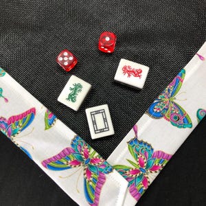 May include: A white fabric game mat with a black mesh center and a white border with a butterfly pattern. There are four red and white dice and four white tiles with Chinese characters and images on them.