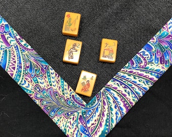 May include: Four rectangular yellow mahjong tiles with red and green illustrations of figures and numbers are arranged on a black mesh surface bordered by a vibrant fabric trim featuring a blue, purple, and green paisley pattern.