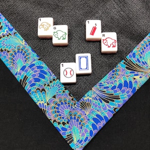 May include: A collection of Mahjong tiles with various symbols, including an eagle, a buffalo, a baseball, and a firecracker. The tiles are arranged on a black surface with a decorative, peacock-feather patterned fabric in shades of blue, purple, and gold.