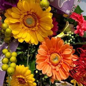 May include: A close-up of a floral wreath with a variety of flowers, including yellow, orange, and pink gerbera daisies. The wreath is decorated with green leaves and purple ribbon.