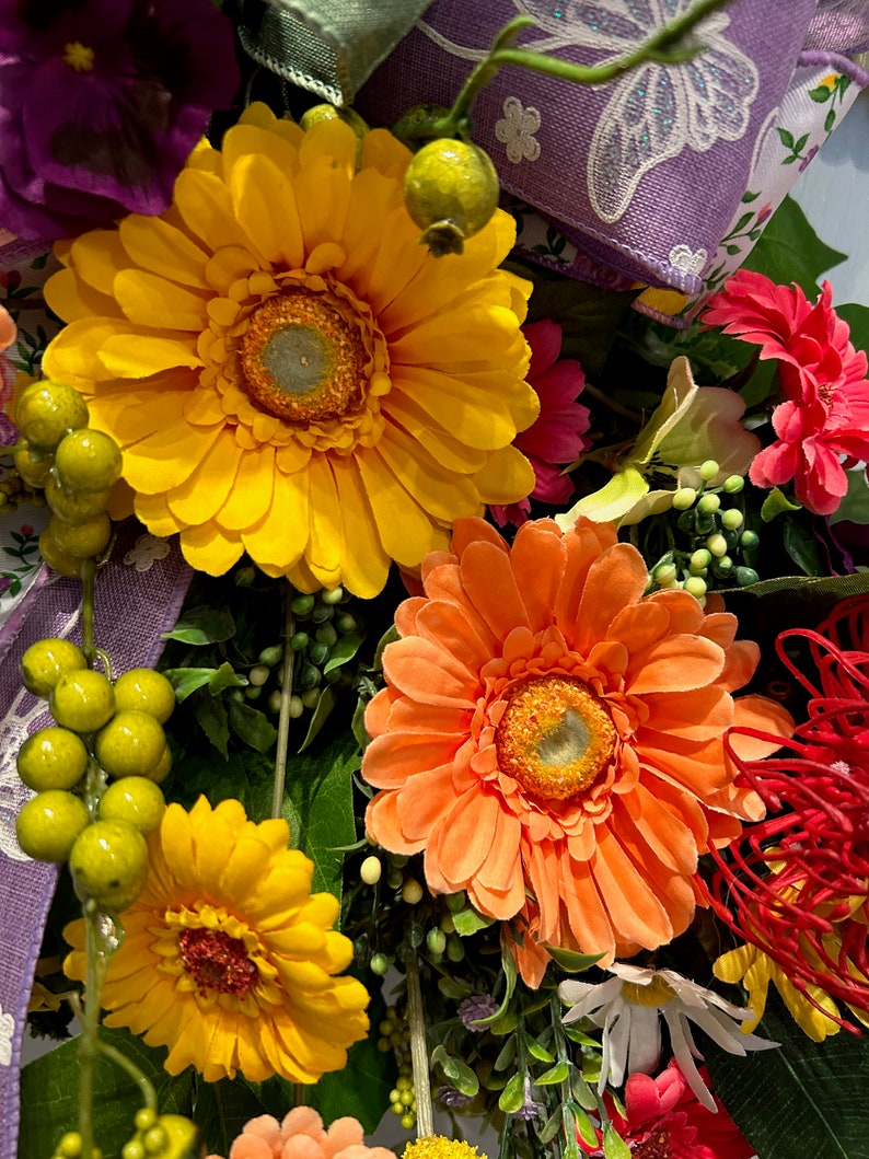May include: A close-up of a floral wreath with a variety of flowers, including yellow, orange, and pink gerbera daisies. The wreath is decorated with green leaves and purple ribbon.