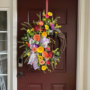May include: A colorful spring wreath with yellow, orange, pink, and purple flowers, green leaves, and a purple and white ribbon hanging on a dark red door.