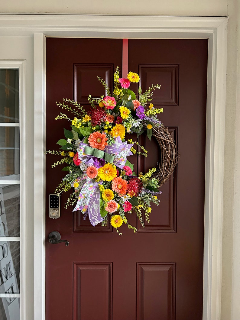 May include: A colorful spring wreath with yellow, orange, pink, and purple flowers, green leaves, and a purple and white ribbon hanging on a dark red door.