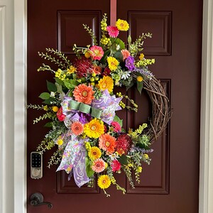 May include: A spring wreath with a variety of colorful flowers, including yellow, pink, purple, and orange. The wreath is made with a grapevine base and has a purple and white ribbon bow.