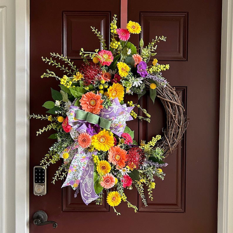 May include: A spring wreath with a variety of colorful flowers, including yellow, pink, purple, and orange. The wreath is made with a grapevine base and has a purple and white ribbon bow.