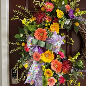 May include: A colorful spring wreath with a variety of flowers, including pink, yellow, orange, and red gerbera daisies, as well as green foliage and a purple and white floral ribbon.
