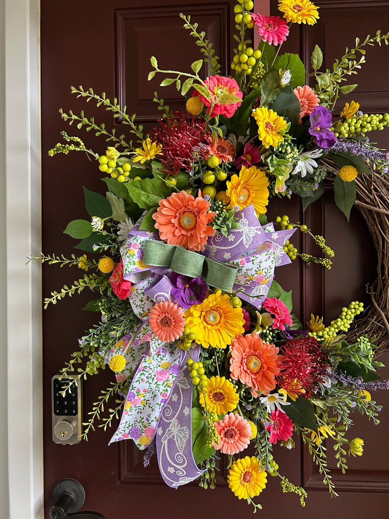May include: A colorful spring wreath with a variety of flowers, including pink, yellow, orange, and red gerbera daisies, as well as green foliage and a purple and white floral ribbon.