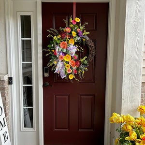 May include: A front door with a wreath made of faux flowers in shades of yellow, orange, pink, and purple. The wreath is hung on a burgundy door with a white door frame. A welcome sign is on the left side of the door and a potted plant with yellow flowers is on the right side of the door.