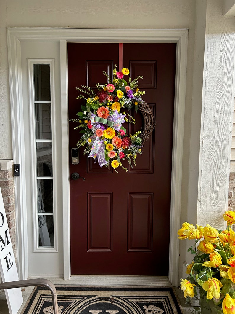 May include: A front door with a wreath made of faux flowers in shades of yellow, orange, pink, and purple. The wreath is hung on a burgundy door with a white door frame. A welcome sign is on the left side of the door and a potted plant with yellow flowers is on the right side of the door.