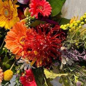 May include: A vibrant floral wreath featuring a variety of flowers, including yellow, orange, pink, and red. The wreath also includes green leaves and a red pinecone-like flower.