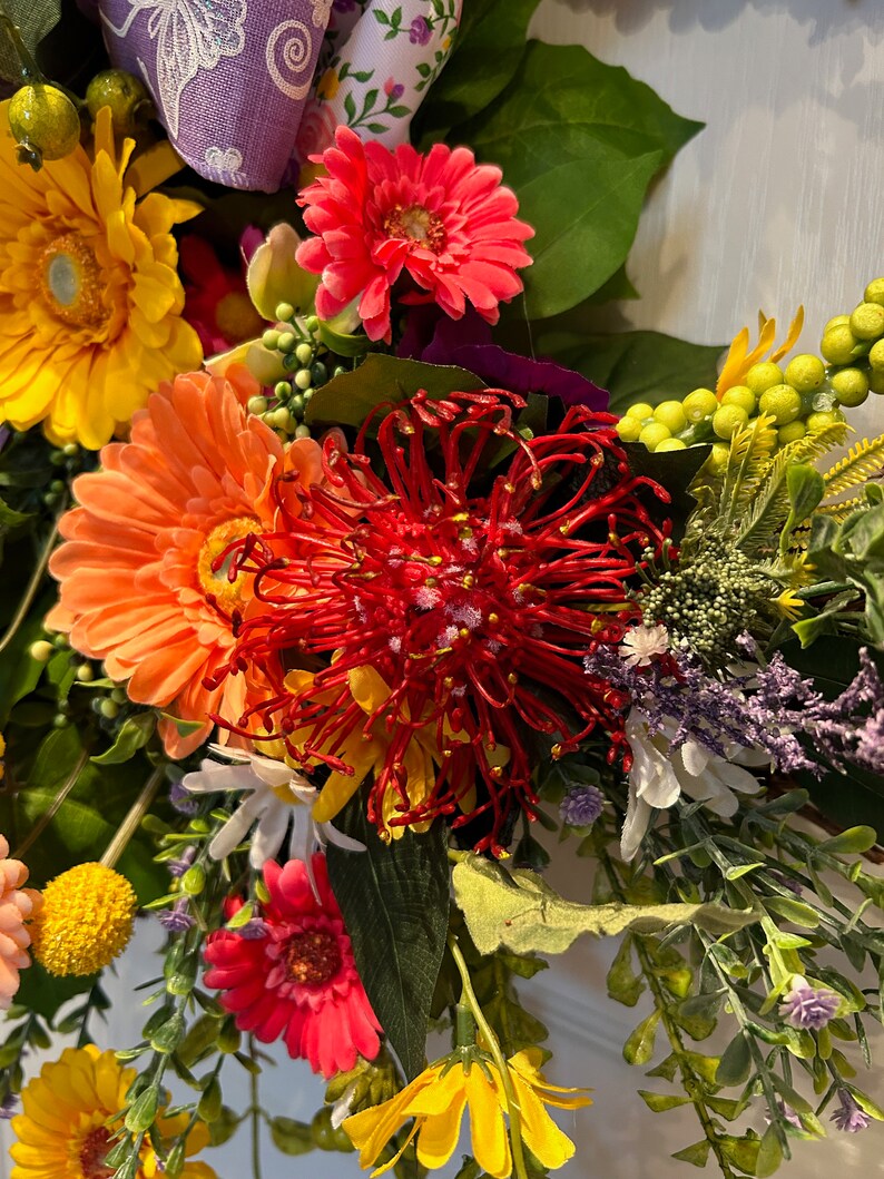 May include: A vibrant floral wreath featuring a variety of flowers, including yellow, orange, pink, and red. The wreath also includes green leaves and a red pinecone-like flower.