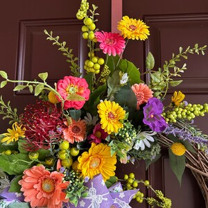May include: A spring wreath with a variety of flowers, including pink, yellow, and orange gerbera daisies, purple pansies, and red pincushion protea. The wreath is decorated with a purple and white ribbon with a butterfly design.
