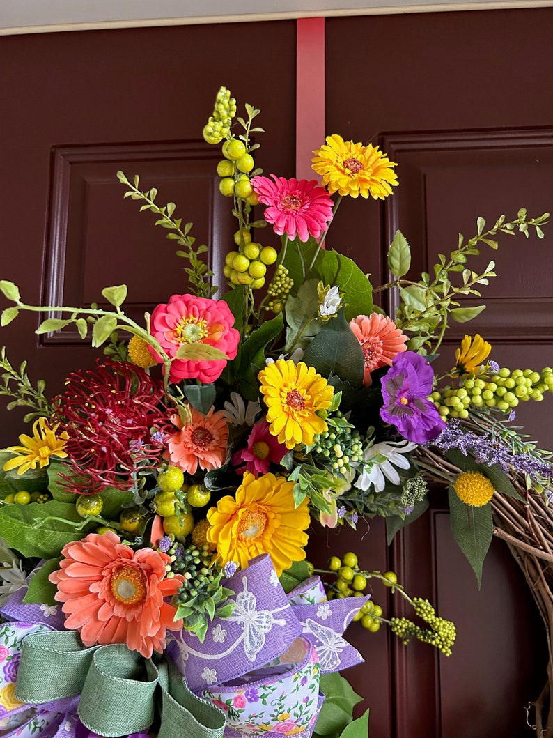 May include: A spring wreath with a variety of flowers, including pink, yellow, and orange gerbera daisies, purple pansies, and red pincushion protea. The wreath is decorated with a purple and white ribbon with a butterfly design.
