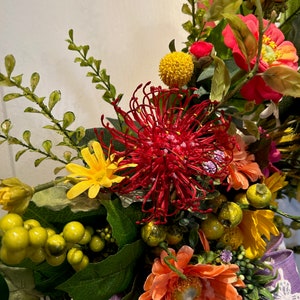 May include: A vibrant floral arrangement featuring a red pincushion protea, orange gerbera daisies, yellow pom poms, and green foliage.