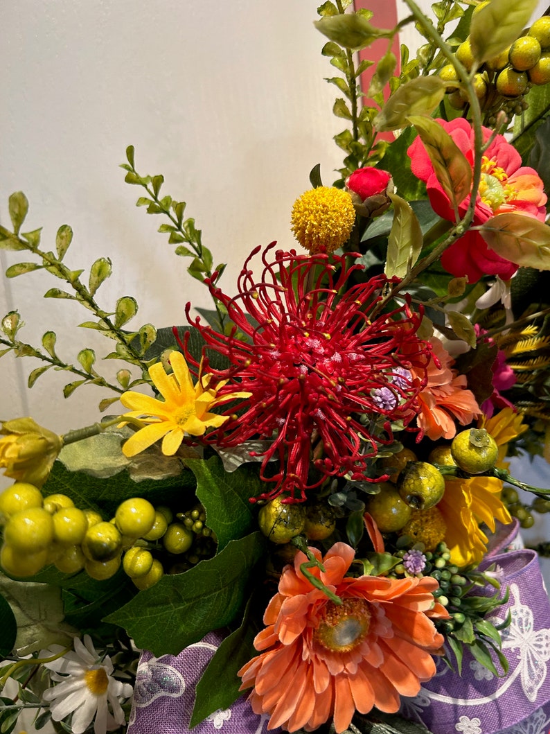 May include: A vibrant floral arrangement featuring a red pincushion protea, orange gerbera daisies, yellow pom poms, and green foliage.
