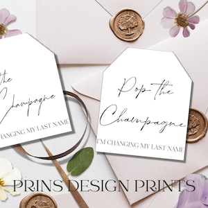Könnte beinhalten: Zwei weiße bedruckbare Geschenkanhänger mit schwarzer Kalligraphie, auf denen "Pop the Champagne I'm Changing My Last Name" steht. Die Anhänger befinden sich auf einem rosa und weißen Hintergrund mit Blumen und Wachssiegeln.