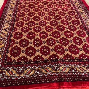 Op de afbeelding: Een rood en goudkleurig tapijt met een franje rand. Het tapijt heeft een traditioneel ontwerp met ingewikkelde details.