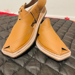 Peut inclure: Une paire de sandales Peshawari en cuir marron clair. Les sandales ont un bout carré et une bride de cheville. Les semelles sont de couleur claire avec une bordure foncée.