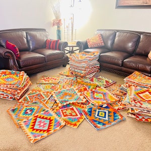 Puede incluir: Una pila de almohadas decorativas de colores con diseños geométricos en tonos rojos, naranjas, amarillos, azules y verdes. Las almohadas están dispuestas sobre una alfombra delante de dos sofás de cuero marrón.