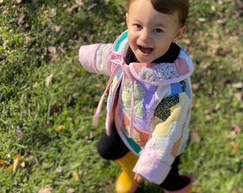 May include: A young child wearing a colourful patchwork jacket and yellow wellies smiles at the camera while standing in a grassy field.