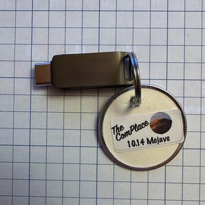 May include: A silver USB-C flash drive with a metal keyring attached to a round tag. The tag has a sticker with the text "The ComPlace 10.14 Mojave" and a landscape image.