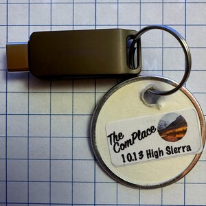May include: A dark olive USB-C adapter with a silver keyring and a round tag. The tag has a sticker with the text "The ComPlace 10.13 High Sierra" and a mountain scene.