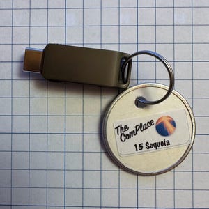 May include: A USB-C connector with a key ring attached to a round metal tag. The tag has a sticker with the text "The ComPlace" and "15 Sequoia" along with a graphic of a blue and orange sphere.