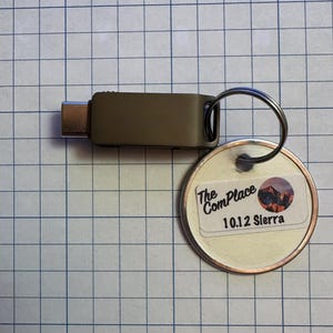 May include: A USB-C flash drive with a key ring attached to a round tag. The tag has a sticker with the text "The ComPlace" and "10.12 Sierra" along with a mountain graphic. The flash drive is olive green.