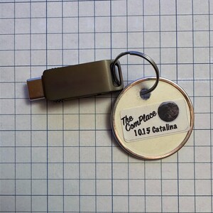 May include: A USB-C flash drive keychain with a silver key ring and a round tag. The tag has a white label with the text "The ComPlace 10.15 Catalina" and a dark circular graphic.