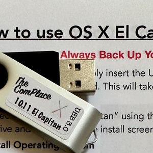 May include: A white USB flash drive with a black cap. The drive is labeled "The Com Place 10.11 El Capitan" and has a USB 3.0 connector. The drive is sitting on a white piece of paper with the text "How to use OS X El Capitan 10.11" and "Always Back Up Your Files Before You" printed on it.